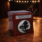 Wooden cremation urn for a pianist with engraved grand piano design, displayed on a stage with a piano in the background.