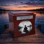 Wooden cremation urn with engraved fisherman silhouette, placed on a lakeside pier at sunset.