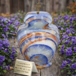 Blue marble cremation urn with gold cross lid, displayed outdoors among purple flowers with memorial plaque.
