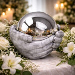 Silver cremation urn shaped like hands holding a reflective sphere, surrounded by white flowers and candles.