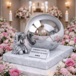 Personalized infant cremation urn with mirrored sphere and grieving angel figure, displayed in a floral memorial setting.