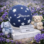 Urna cineraria azzurra per bambini con stelle e nome, esposta con orsetti di peluche in un campo di fiori.