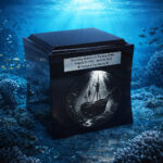 Black cremation urn with engraved vintage ship design, displayed underwater among coral reef and ocean life.