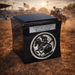Black cremation urn with engraved tribute and rodeo cowboy design, placed in a dusty arena during a rodeo event with an American flag in the background.