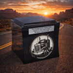 Black cremation urn with engraved tribute and semi-truck design, placed on an open highway at sunset in a desert landscape.