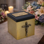 Gold cremation urn with crucifix detail and black lid, placed on a table with candles and floral arrangements in a memorial setting.