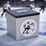 Stainless steel cremation urn with wooden lid and engraved ice hockey player design, placed on an ice rink during a game.