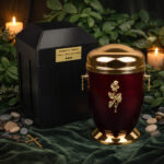 Red and gold cremation urn with rose detail beside a black urn vault, displayed with candles and greenery.