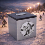 Stainless steel cremation urn with wooden lid and engraved snowboarder design, placed on a snowy ski slope with people in the background.