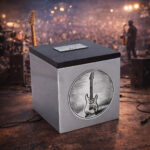 Stainless steel cremation urn with wooden lid and engraved electric guitar design, displayed on a concert stage with musicians and crowd in the background.