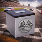Stainless steel cremation urn with wooden lid and engraved racing flags design, placed on a racetrack with cars in motion.