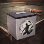 Stainless steel cremation urn with wooden lid and engraved table tennis player design, placed beside a ping pong table with paddles and ball.
