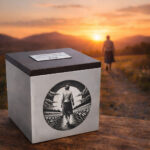 Stainless steel cremation urn with wooden lid and engraved woman walking along a path, set against a peaceful sunset landscape.