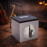 Stainless steel cremation urn with wooden lid and engraved saxophone design, placed on a stage with musical instruments in the background.
