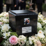 Elegant black cremation urn displayed at a funeral ceremony, surrounded by soft white and pink flowers with a small memorial photo attached.