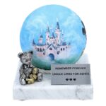 Blue memorial urn for child with castle artwork and teddy bear ornament on marble stand.