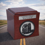 Wooden memorial urn with a truck engraving placed on a rural road with open fields in the background.