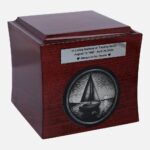 Urna cineraria de madera con velero grabado y placa conmemorativa, vista frontal.